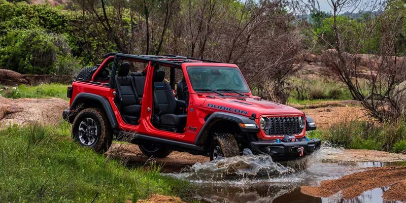 A red Jeep Rubicon, with doors removed, drives through a shallow stream in a natural, rocky setting.