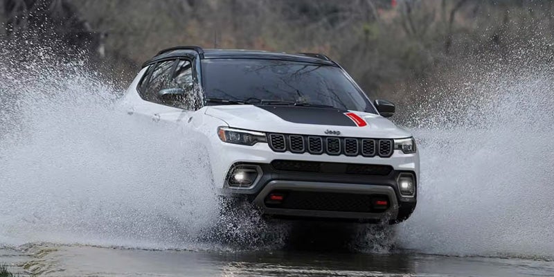 A white Jeep Compass with a black hood stripe and red accents drives through a large puddle, splashing water.