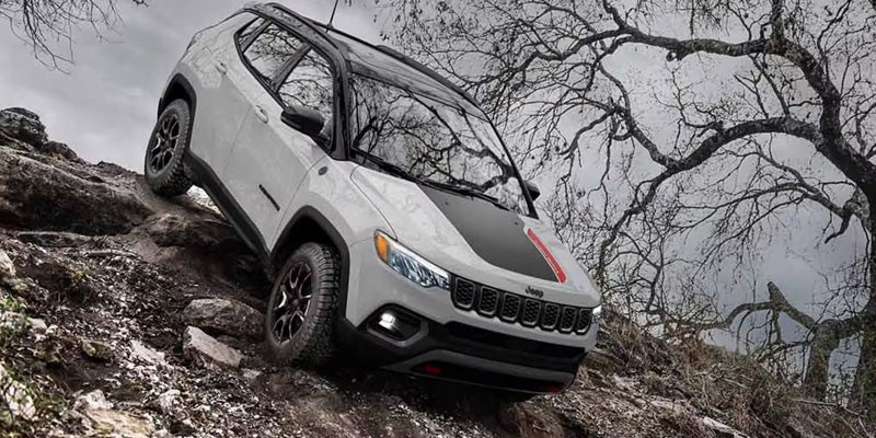 White Jeep Compass with a black hood and red stripes climbing a rocky hill with bare trees in the background.