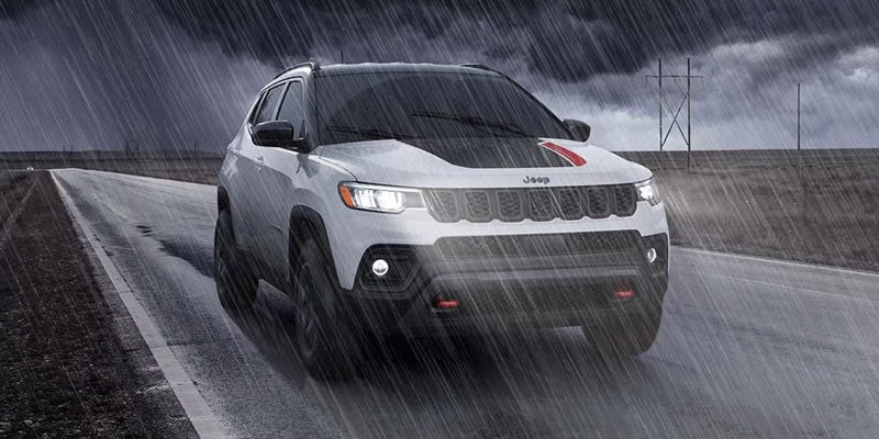 White Jeep Compass Trailhawk driving on a rain-soaked road with headlights illuminated