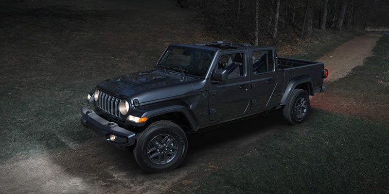 Dark-colored Jeep Gladiator driving on a dirt path at night, illuminated by headlights