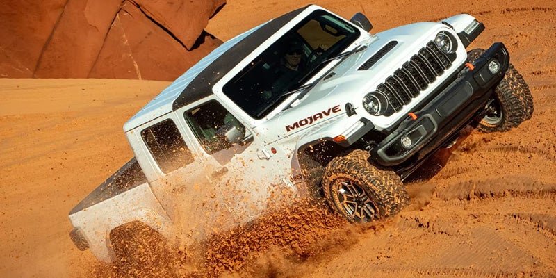 White Jeep Gladiator Mojave driving through deep sand, kicking up dust while climbing a desert dune.