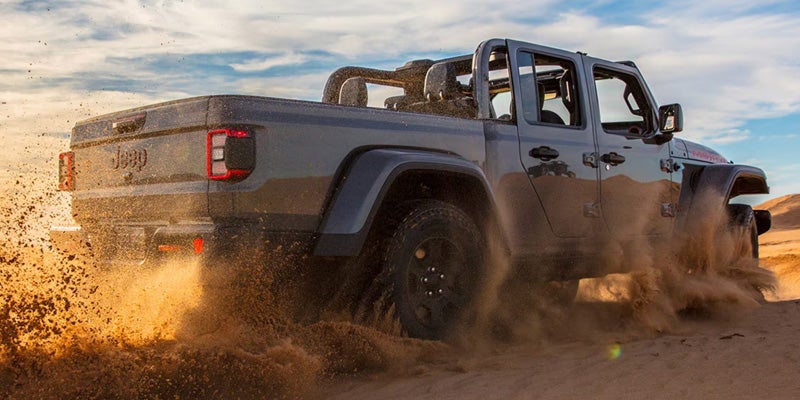 Gray Jeep Gladiator driving through sand with dirt spraying behind the vehicle, showcasing the pickup bed