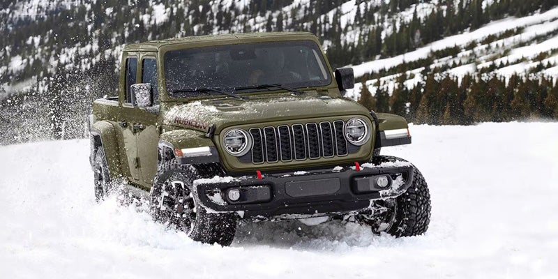 Green Jeep Gladiator driving through deep snow, with snow spraying around the tires in a mountain landscape.