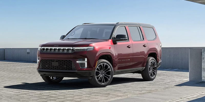 A deep red Jeep Grand Wagoneer SUV is parked on a paved lot with a concrete wall in the background.