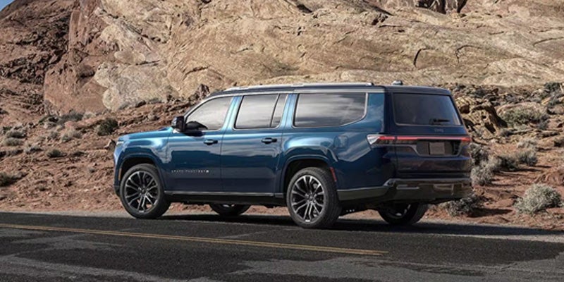 A dark blue 2023 Grand Wagoneer L SUV parked on a paved road with a rugged, rocky mountain backdrop.