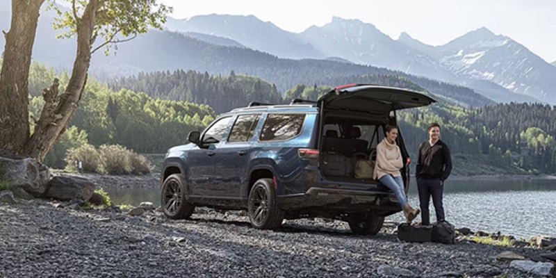 Blue Jeep Wagoneer parked by a lakeside with the rear liftgate open, highlighting cargo space against a mountain backdrop.