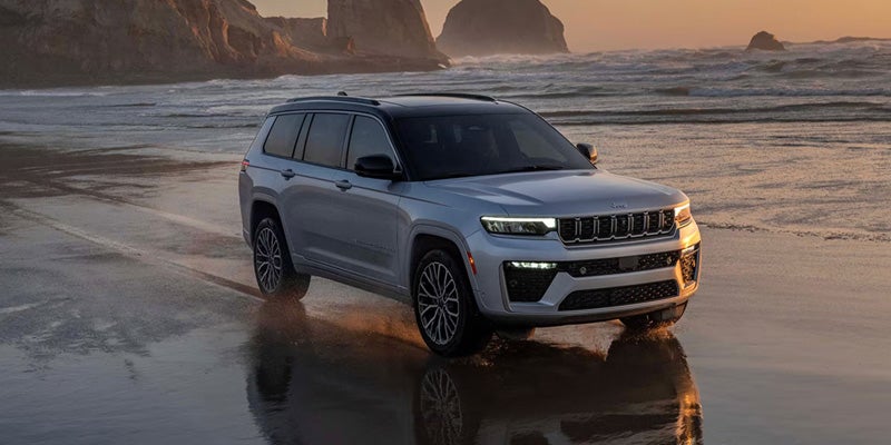 Silver SUV driving on a wet sandy beach near breaking waves and large rock formations under a sunset sky.