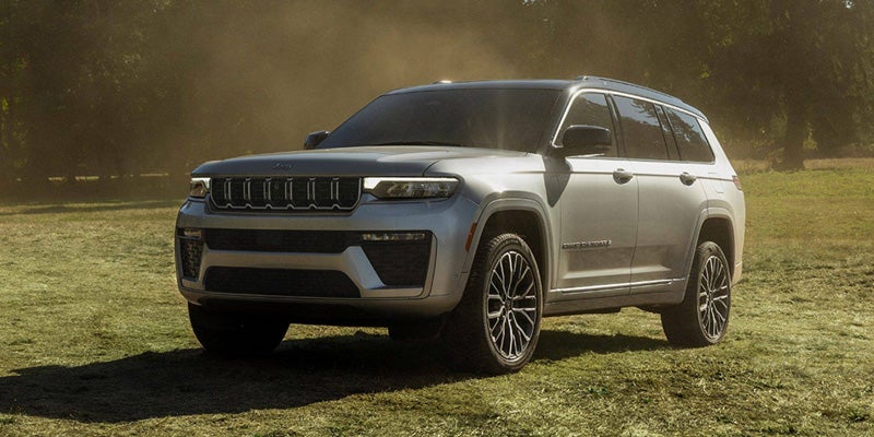 A silver Jeep Grand Cherokee L, with a black roof, parked on a grassy field with trees in the background.