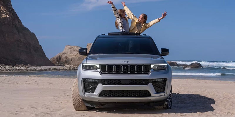 White Jeep Grand Wagoneer parked on a beach with passengers standing through the open sunroof