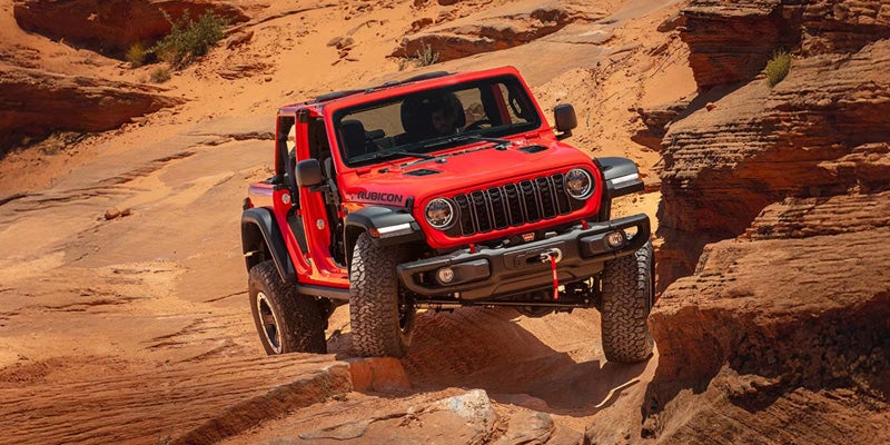 Red off-road SUV driving over rocky desert terrain with rugged sandstone cliffs in the background.