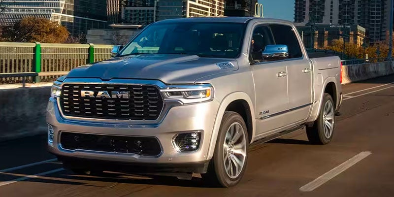 A silver Ram 1500 pickup truck drives on a city highway with buildings in the background.