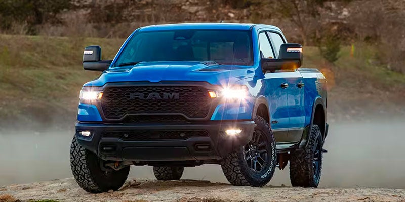 Front view of a blue Ram 1500 TRX pickup truck with headlights on, parked on a dirt road.
