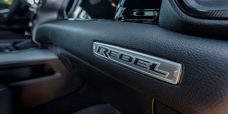 Close-up of a silver "REBEL" emblem with black lettering embedded in a textured black car dashboard.
