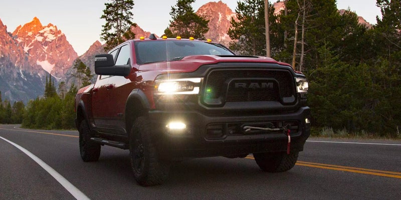 Front view of a red Ram truck with headlights on, driving on a road with mountains and trees in the background.