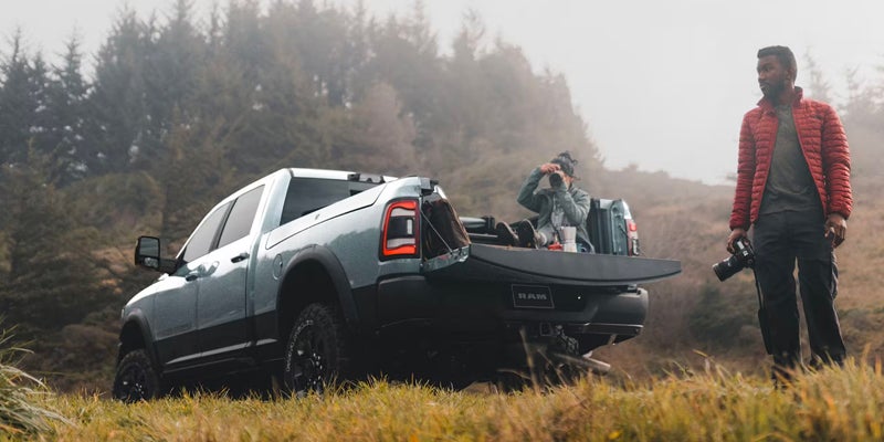 A light blue Ram truck parked on a grassy hill with its tailgate down. One person is sitting on the tailgate taking a photo, while another stands nearby holding a camera.