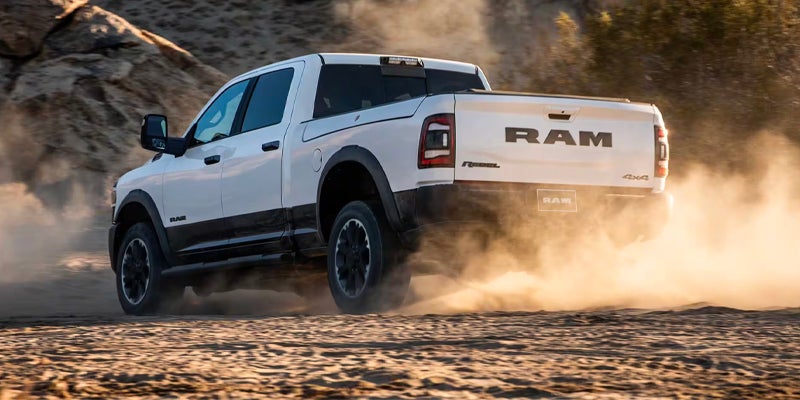 Rear view of a white Ram Rebel pickup truck kicking up dust as it speeds across sandy terrain.