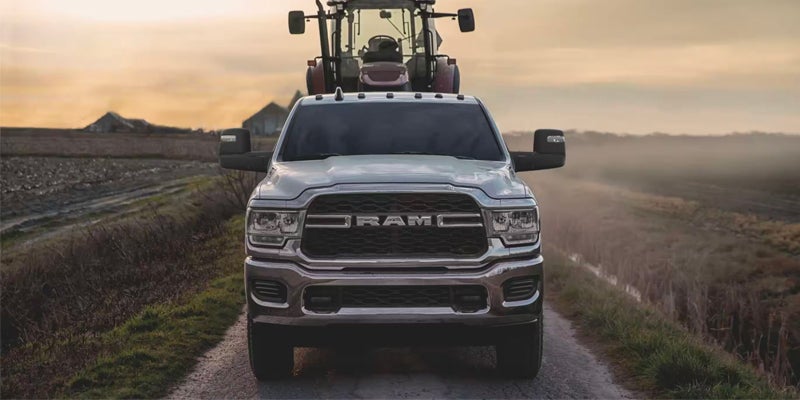 A white RAM pickup truck faces forward on a gravel road, with a red tractor behind it, framed by fields at dusk.