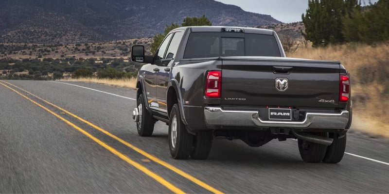Rear view of a dark gray Ram 3500 dually truck driving on a two-lane road through arid hills.