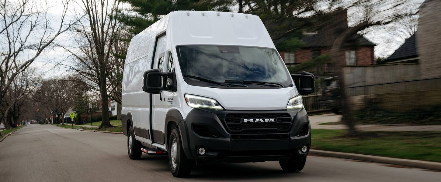 A white 2023 Ram ProMaster van driving on a residential street with bare trees and houses in the background.