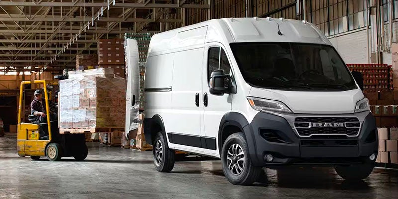 A white Ram ProMaster van with its side door open, and a forklift operator loading boxes into it in a warehouse.