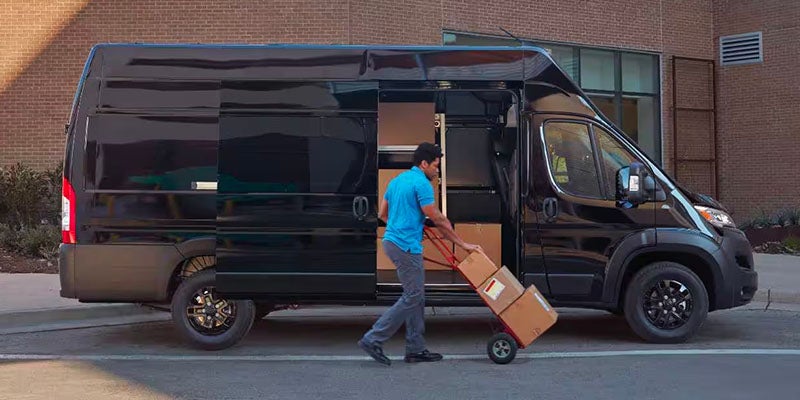 A man in a blue shirt and grey pants loads boxes on a hand truck into an open black delivery van.