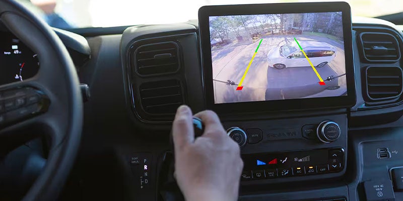 Hand shifting gears in a vehicle with a backup camera displaying a white car and colored parking lines.
