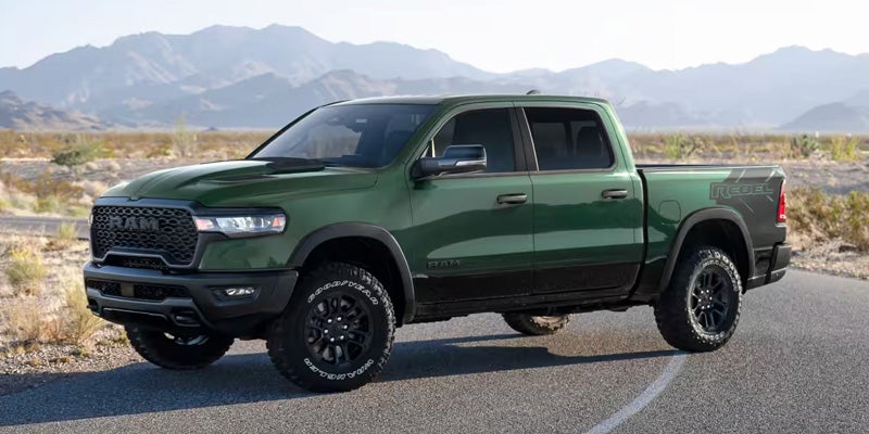 A green 2023 Ram 1500 Rebel pickup truck, angled right, on an asphalt road with mountains in the background.