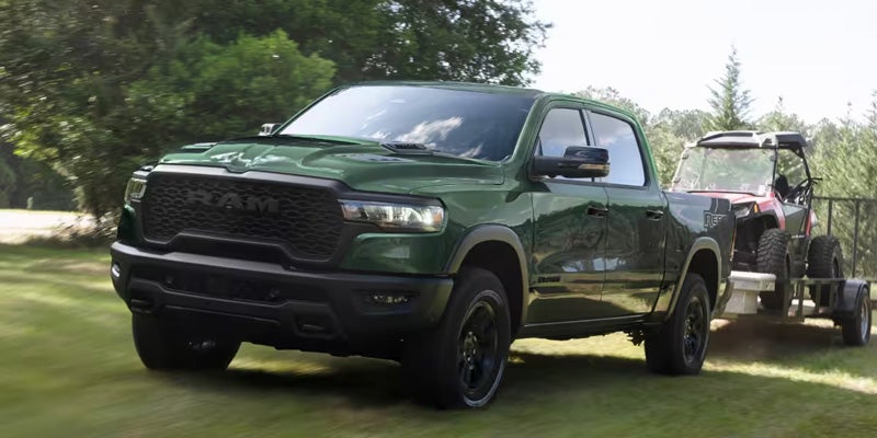 A green Ram TRX pickup truck towing a red off-road vehicle on a trailer, driving on green grass.