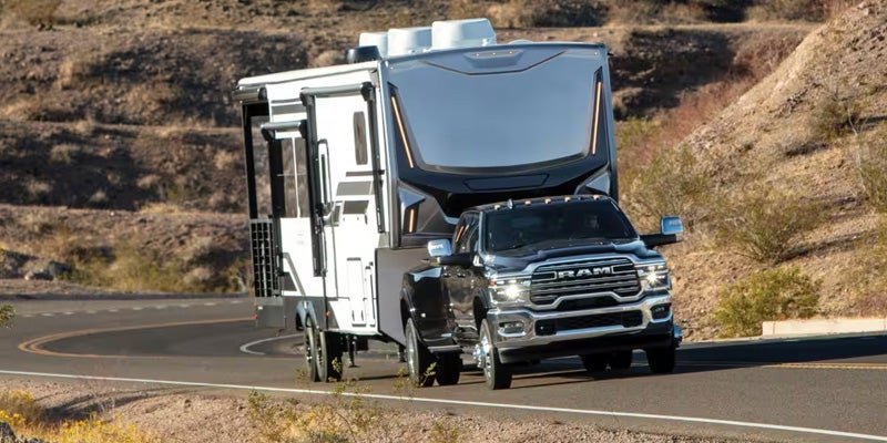 A black Ram 3500 pickup truck towing a white fifth-wheel RV on a winding desert road.