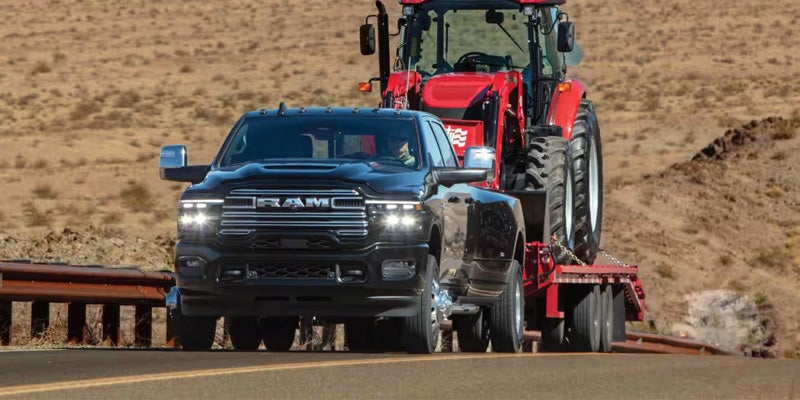 A black Ram 3500 pickup truck tows a red tractor on a flatbed trailer down a desert highway.