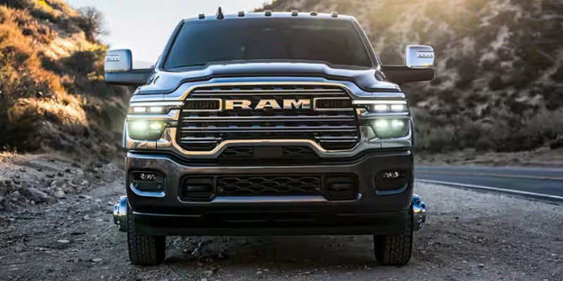 Front view of a dark gray Ram pickup truck with headlights on, parked on a dirt road next to a highway.