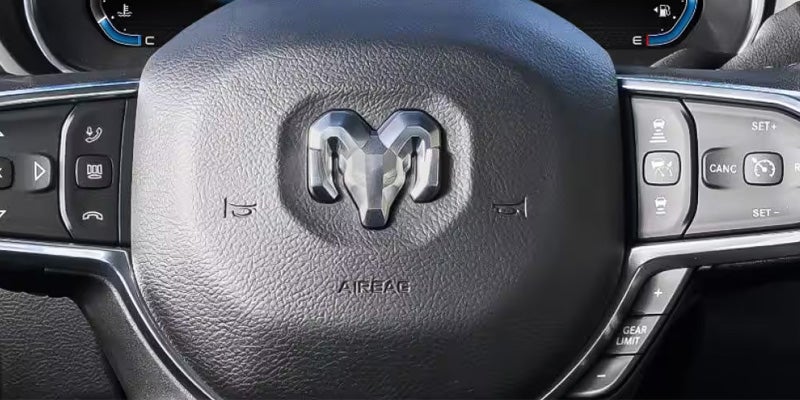A close-up of a Ram truck steering wheel showing the silver Ram logo, control buttons, and the word "AIRBAG".