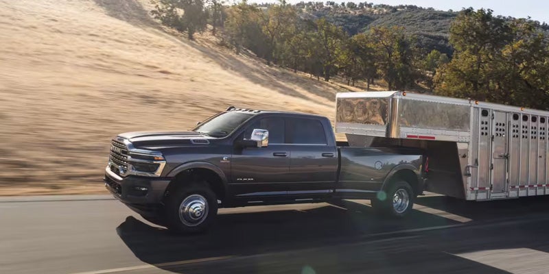 A gray Dodge Ram dually truck pulls a large silver livestock trailer down a highway with hills and trees in the background.