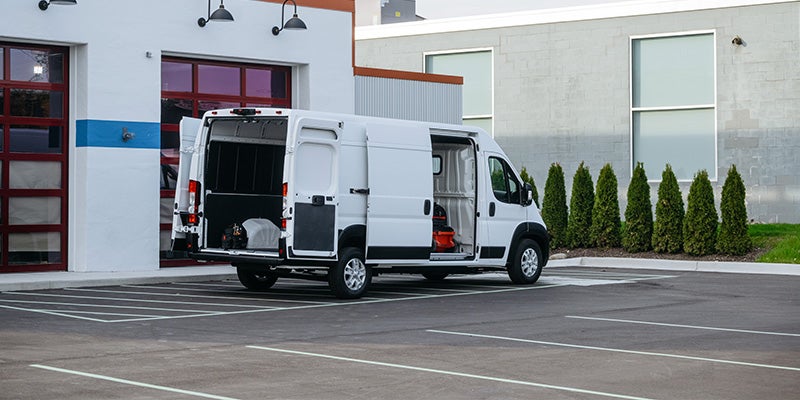 White cargo van with all doors open, parked in a lot in front of a white building with red garage doors.
