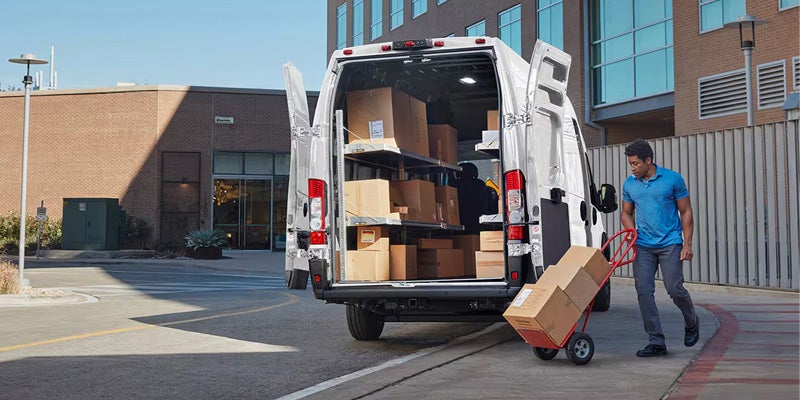 A delivery driver uses a hand truck to unload boxes from the back of a white cargo van.