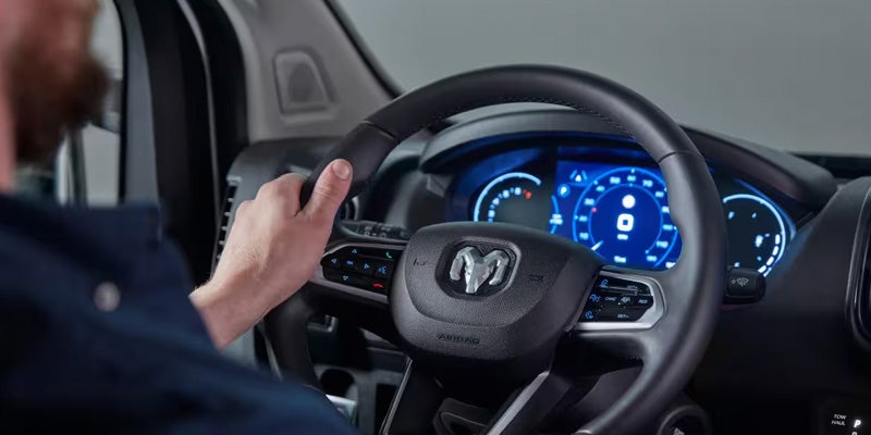 Driver's hand on a black steering wheel with a Ram logo and blue digital dashboard in the background.