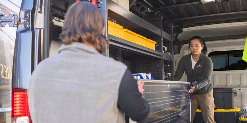 Two people loading a solar panel into the back of a cargo van with shelving and yellow storage bins.