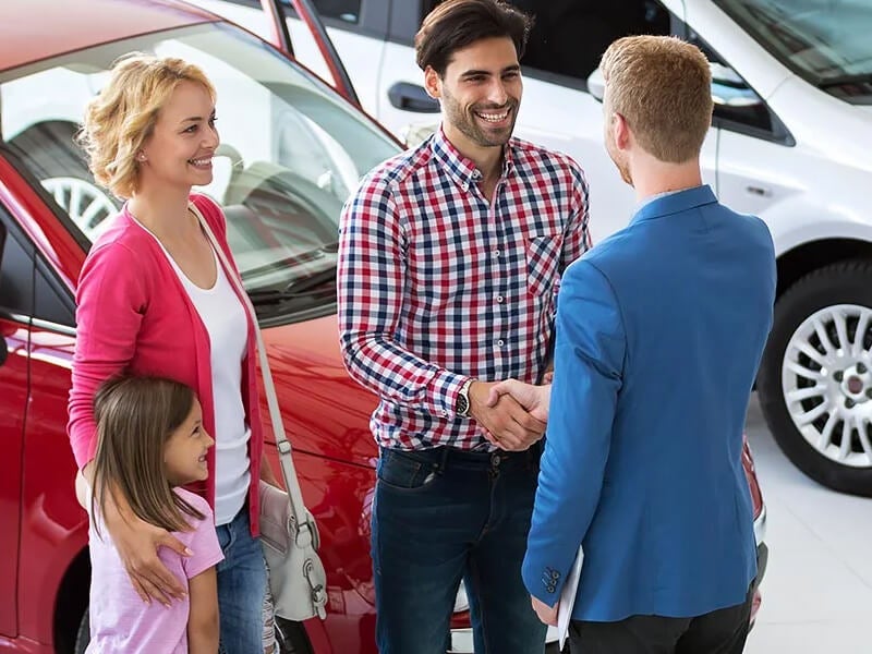 A family, with a smiling mother and daughter, watches as a father shakes hands with a car salesman at a dealership.