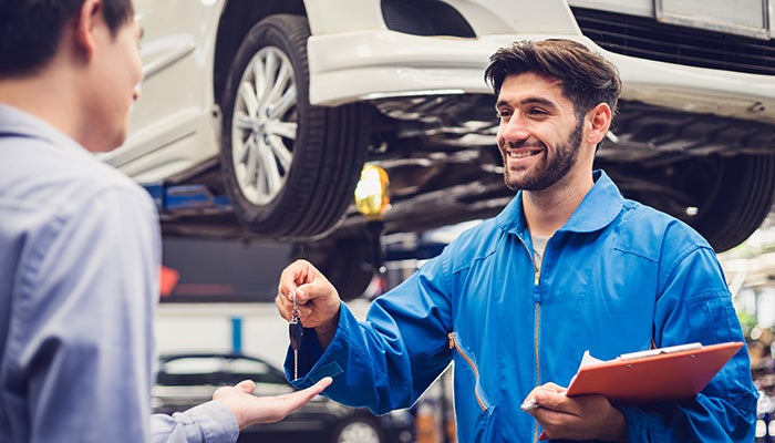 Smiling mechanic in blue coveralls hands car keys to a customer in a repair shop with a car on a lift.